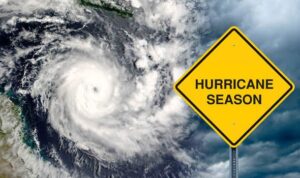 A swirling hurricane is shown in satellite view beside a yellow road sign warning of "Hurricane Season" under a cloudy sky.