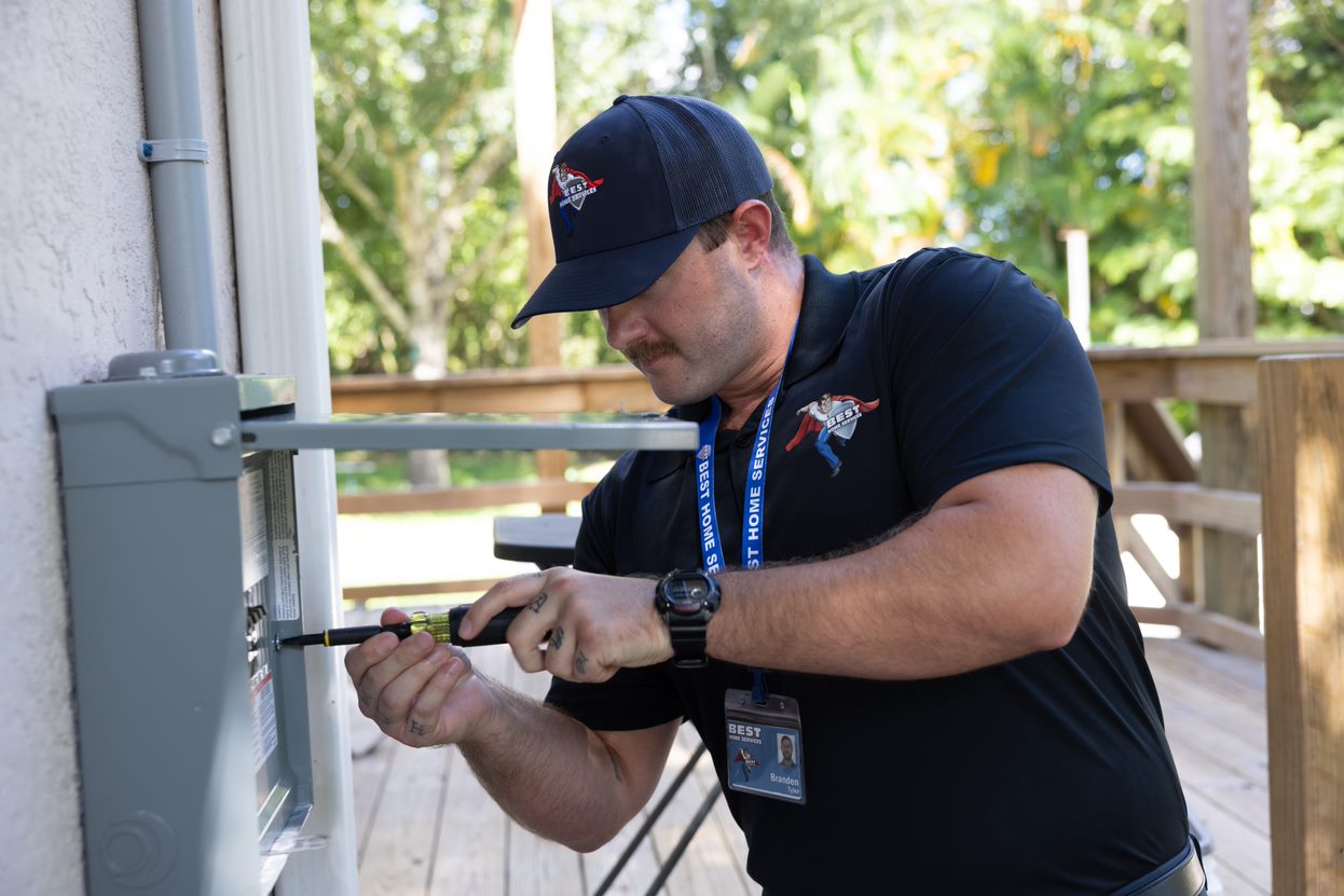 A man in a cap uses a screwdriver on an outdoor electrical panel, wearing a lanyard and branded polo shirt. Trees and a wooden structure are in the background.