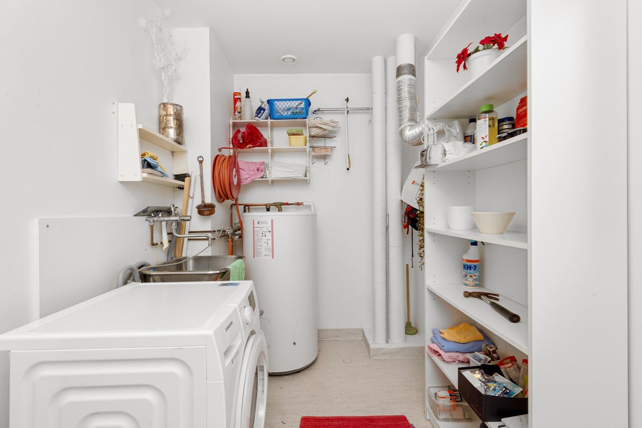 A utility room with a washing machine, sink, water heater, and shelves holding cleaning supplies, towels, and various household items.